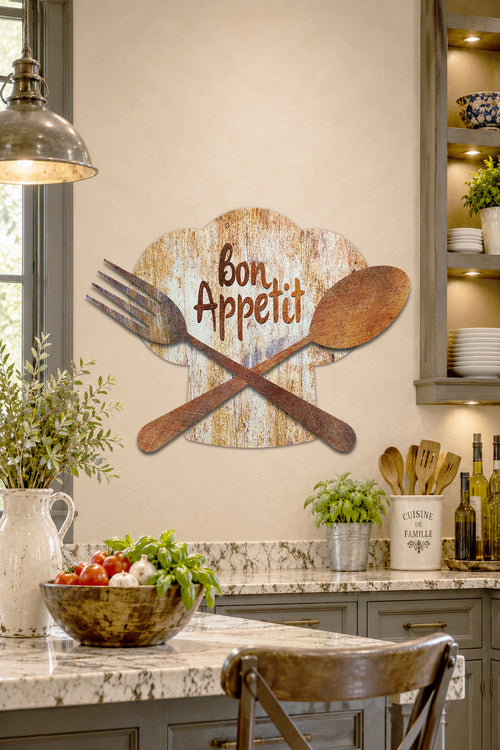 A rustic wall sign reading 'Bon Appetit' in brown lettering, mounted on a beige kitchen wall above a marble countertop. The sign features a distressed wood-textured cloud shape with a crossed wooden spoon and fork in front.
