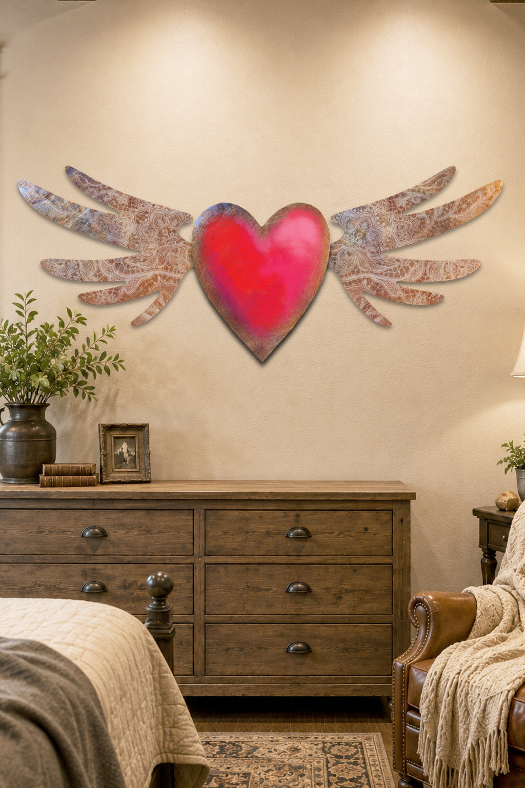 A decorative wall art piece featuring a bright red heart with distressed silver wings, mounted on a beige wall above a wooden dresser in a styled bedroom setting.