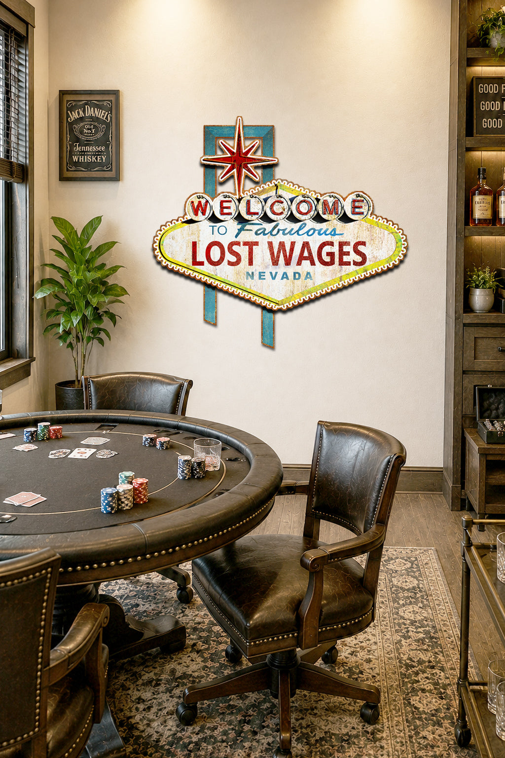A decorative wall sign reading 'Welcome to Fabulous Lost Wages Nevada' mounted on a beige wall in a furnished room with a round poker table, dark leather chairs, shelving, and a plant.